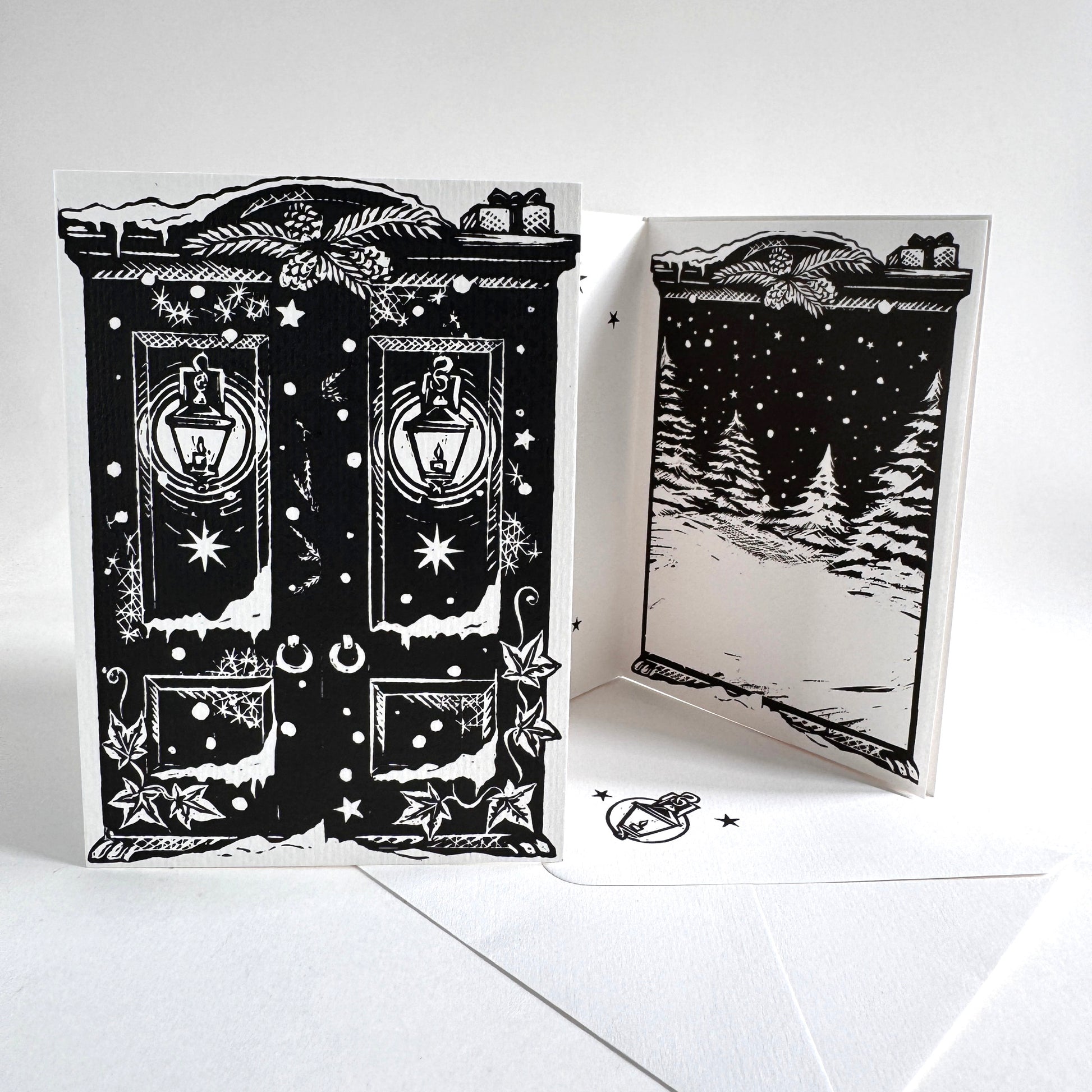 A black and white illustrated greeting card set shown on a white background, with one card standing flat and another opened to show a snowy night scene. The card design features lanterns, stars, snowflakes, and decorative border details.