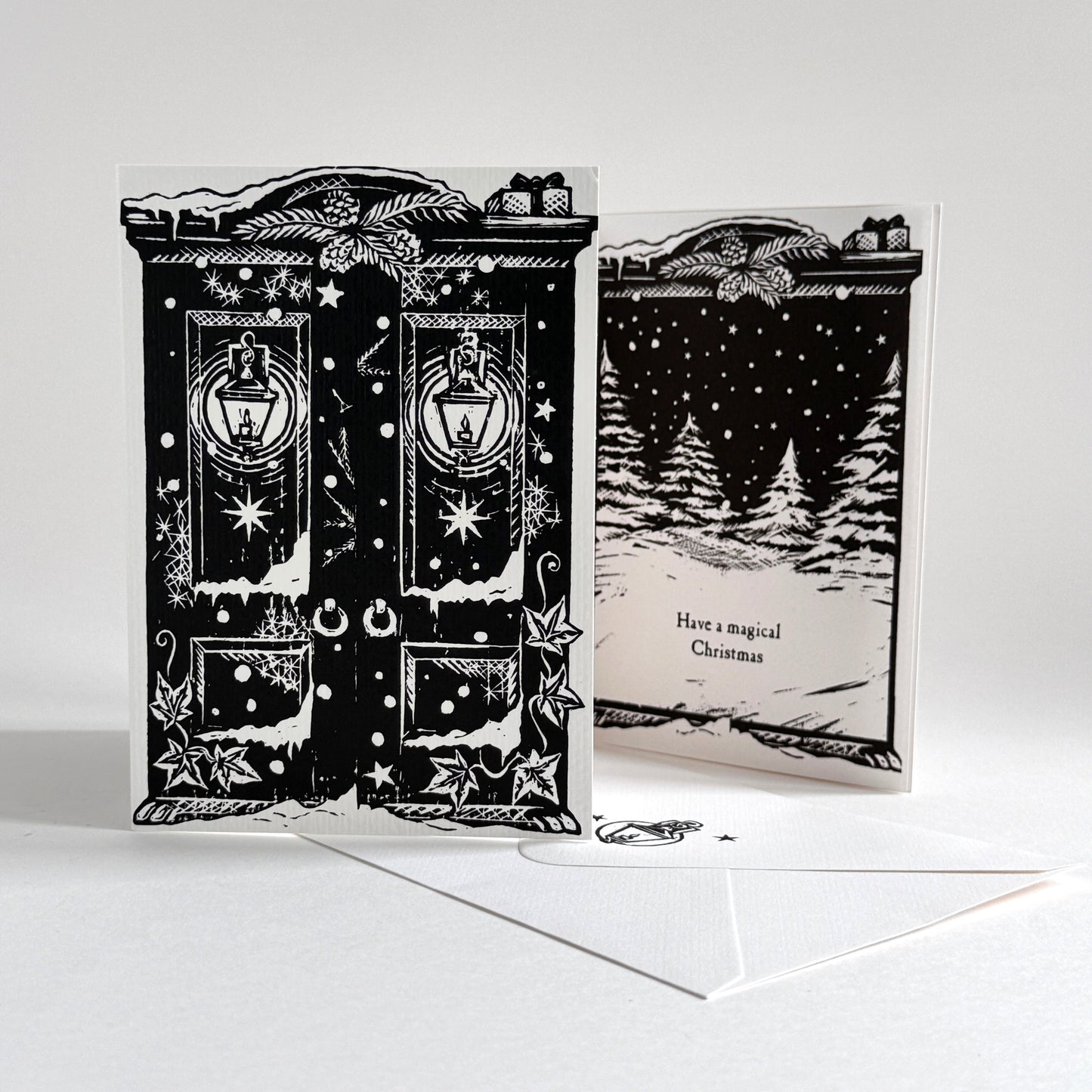 A black and white double-sided Christmas greeting card set with an envelope. The front features an ornate double door design with lanterns and stars, and the back shows a snowy forest scene with the text 'Have a magical Christmas'.