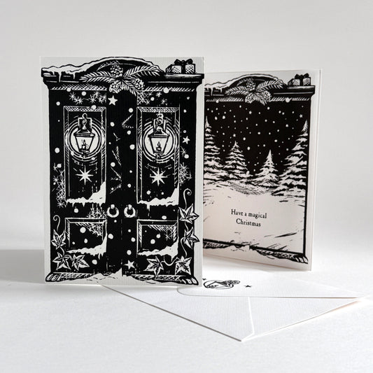 A black and white double-sided Christmas greeting card set with an envelope. The front features an ornate double door design with lanterns and stars, and the back shows a snowy forest scene with the text 'Have a magical Christmas'.