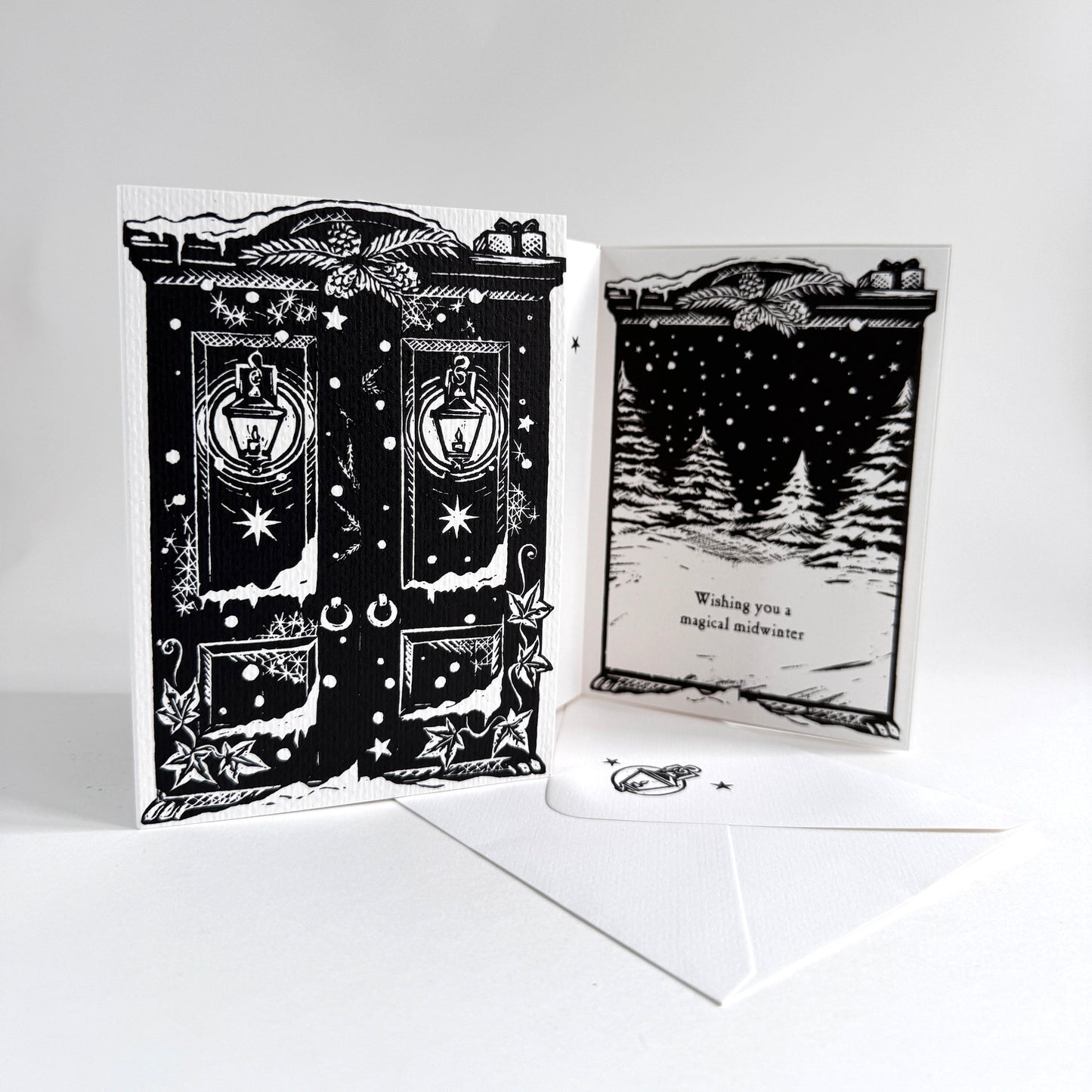 A black and white illustrated greeting card set displayed on a white surface against a light gray background. The set includes a front panel showing a detailed black-and-white door illustration with lanterns and stars, a back panel with a snowy tree scene and the text 'Wishing you a magical midwinter' and a white envelope.