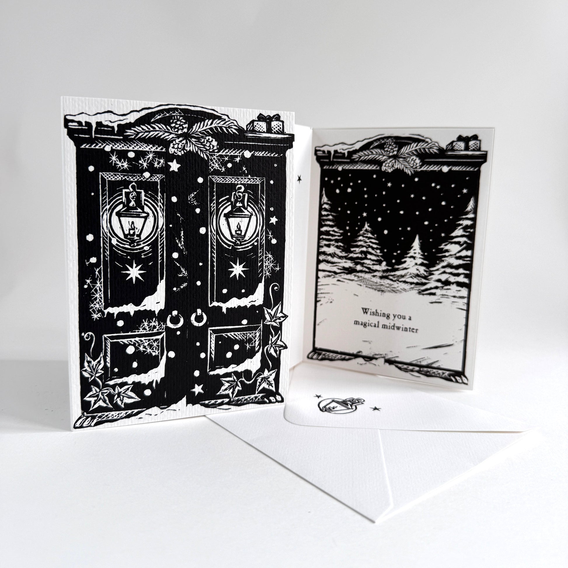 A black and white illustrated greeting card set displayed on a white surface against a light gray background. The set includes a front panel showing a detailed black-and-white door illustration with lanterns and stars, a back panel with a snowy tree scene and the text 'Wishing you a magical midwinter' and a white envelope.