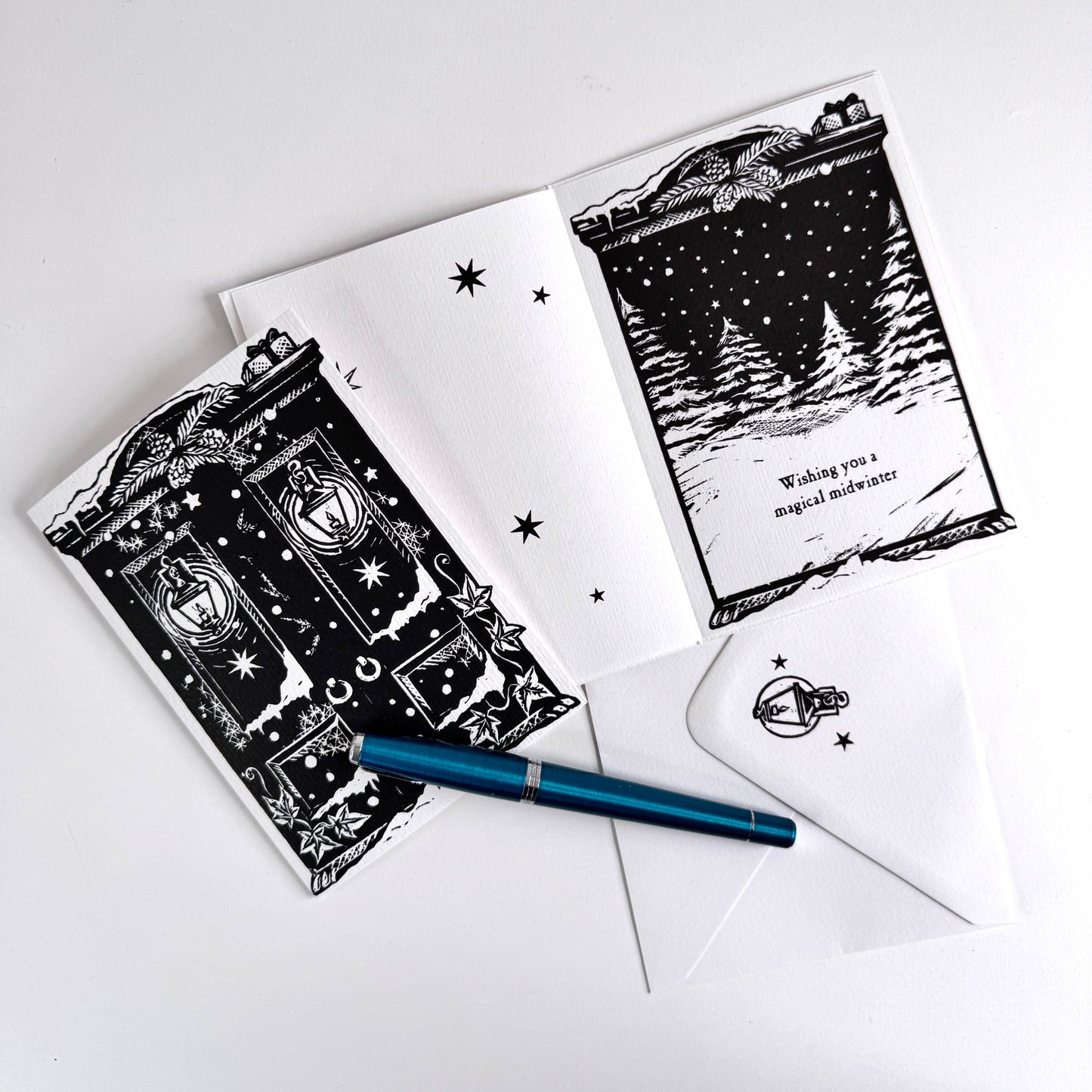 A flat-lay of a black and white illustrated greeting card set with an open card showing a snowy house scene and text, a matching closed card, a white envelope, and a blue pen on a light gray background.
