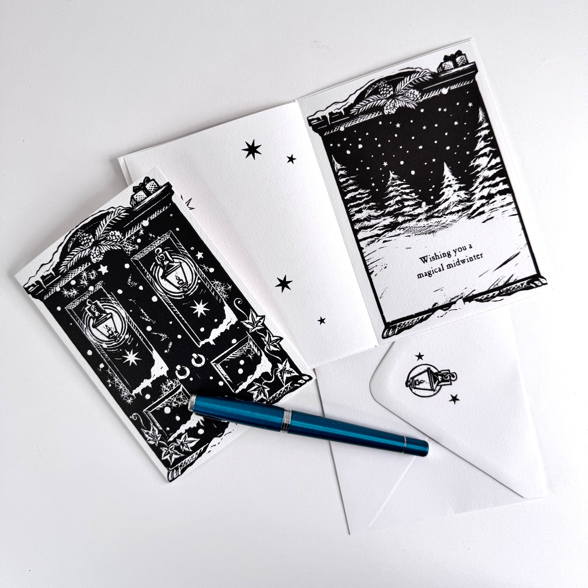 A flat-lay of a black and white illustrated greeting card set with an open card showing a snowy house scene and text, a matching closed card, a white envelope, and a blue pen on a light gray background.