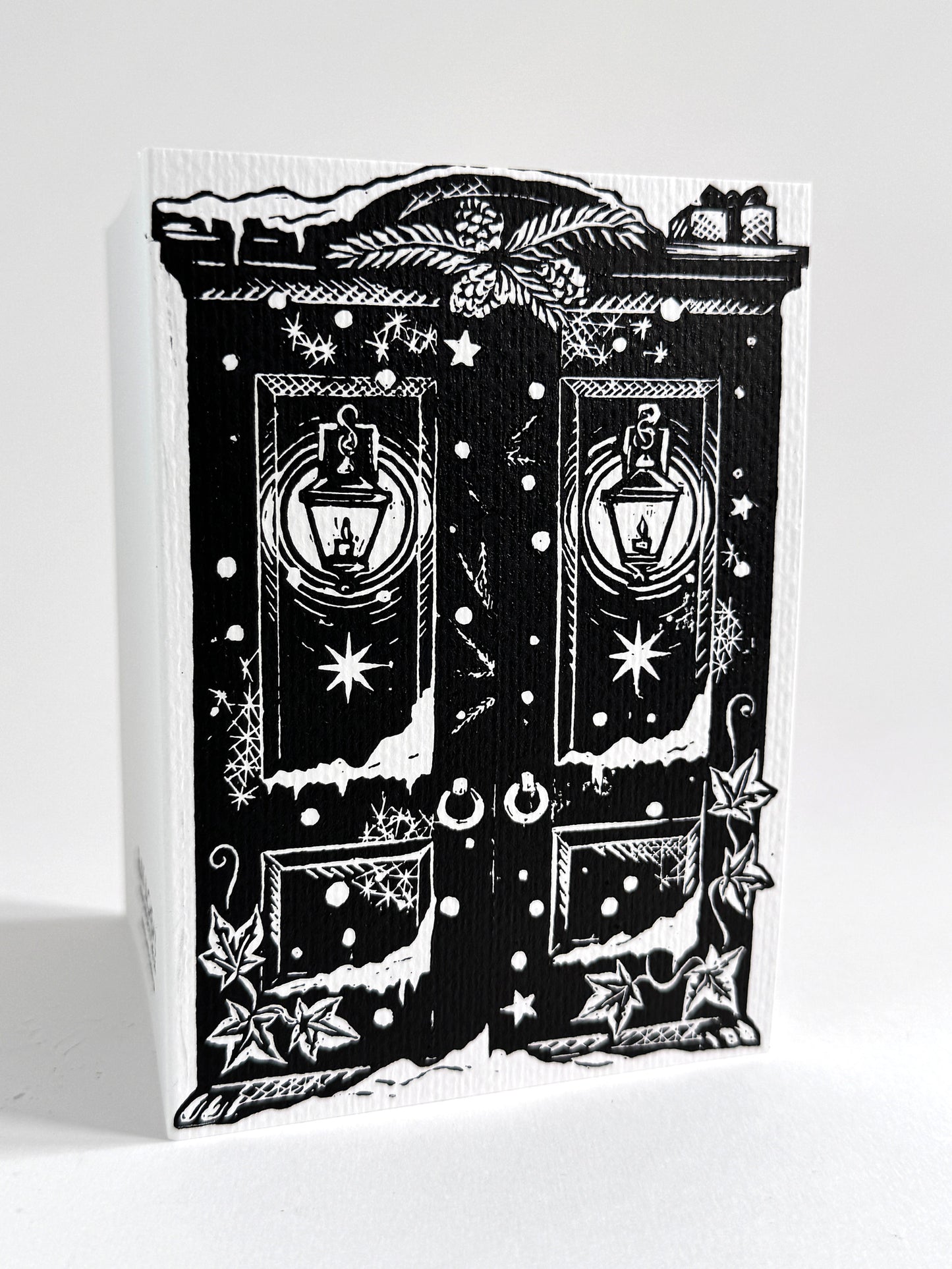 A black and white linoprint greeting card featuring a whimsical double-door design with lanterns, stars, snow-like marks, and decorative motifs, photographed on a white background.