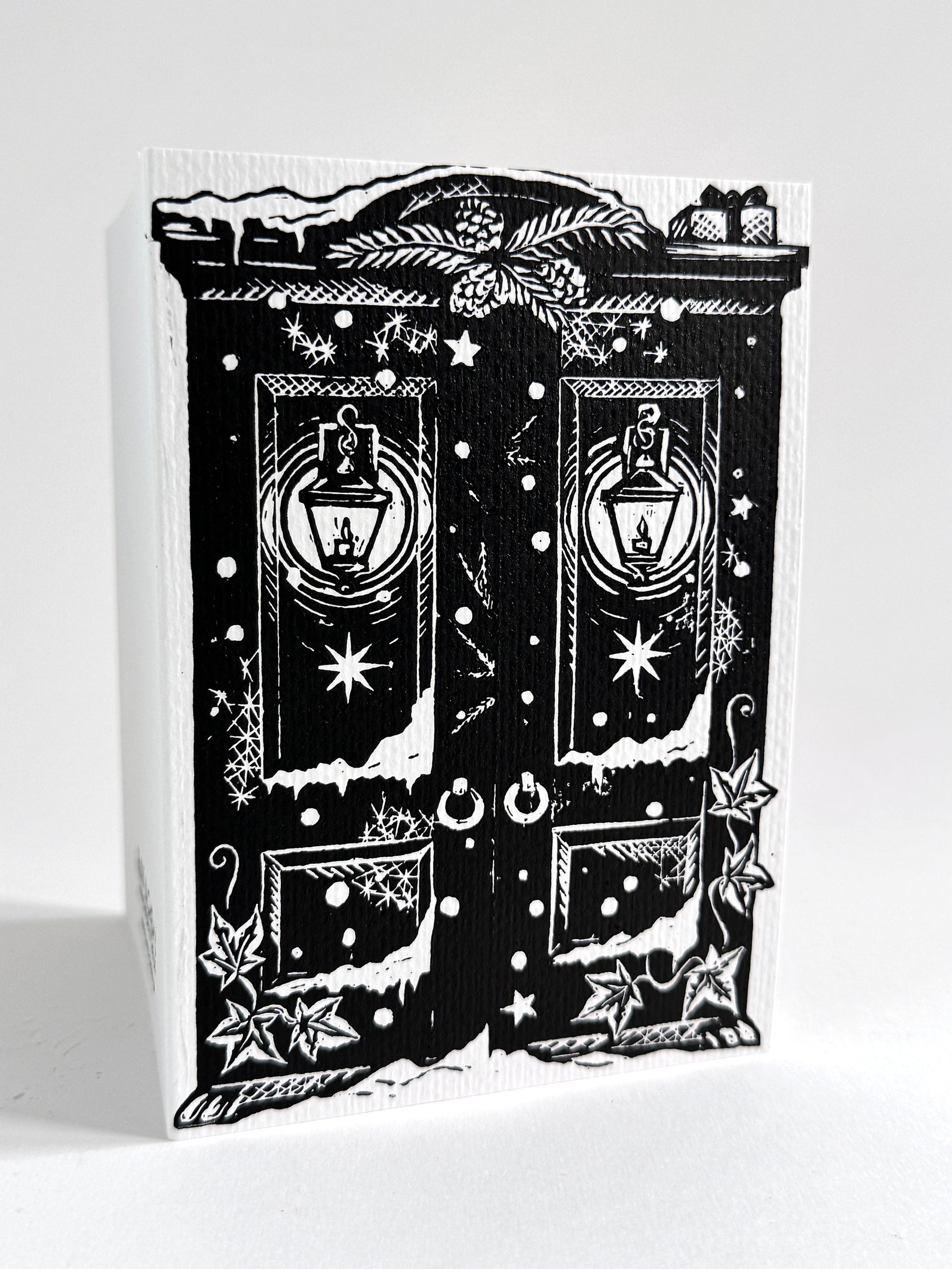 A black and white linoprint greeting card featuring a whimsical double-door design with lanterns, stars, snow-like marks, and decorative motifs, photographed on a white background.
