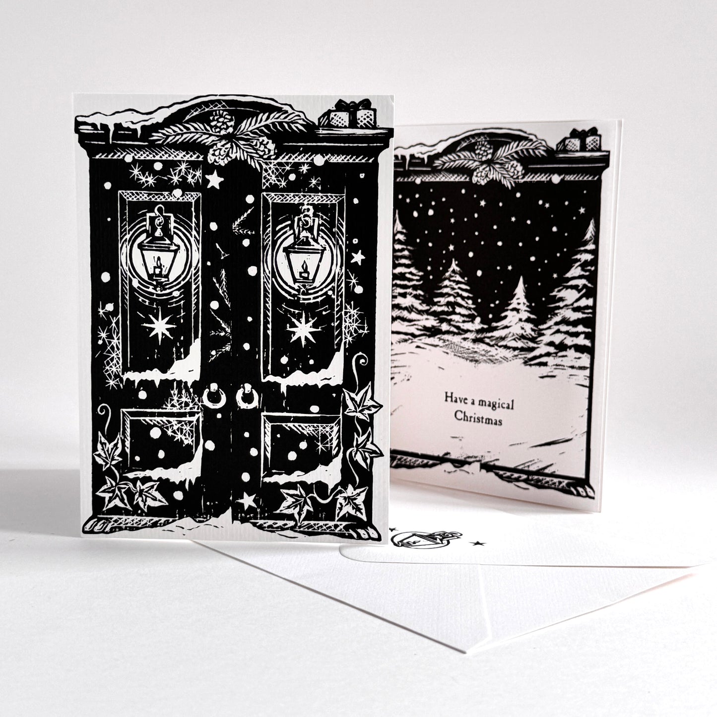 A black and white Christmas-themed greeting card set displayed on a white surface. The front shows two ornate snow-covered doors with lanterns and stars, and the inside shows a snowy forest scene with the text 'Have a magical Christmas.