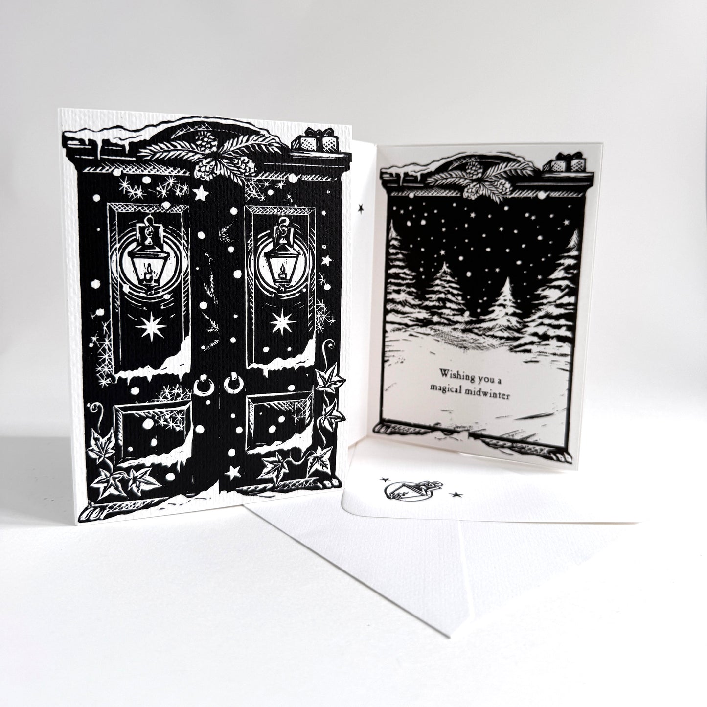 A black and white double-sided greeting card and envelope set. The front features an intricate illustration of a Christmas wardrobe with lanterns, stars, and holly, while the back shows a snowy pine tree scene with the text 'Wishing you a magical midwinter'.