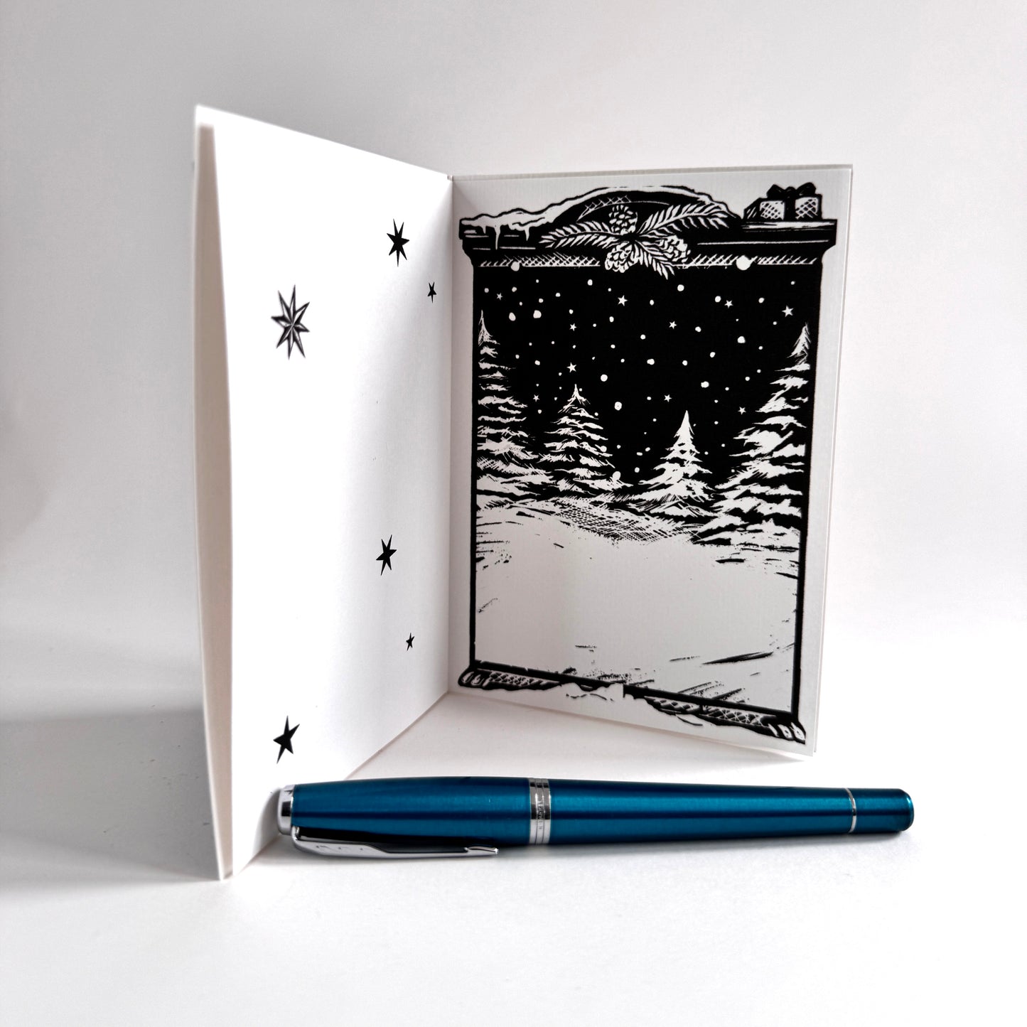 A black and white woodcut-style greeting card shown open, with a snowy forest and stars printed on the inside. A metallic teal pen lies horizontally in front of the card on a clean white background.